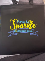Load image into Gallery viewer, A black fabric tote bag with the text 'Born to Sparkle RHOSEBUD CLUB' printed on the front in yellow, blue, and white, resting on a gray fabric surface.

